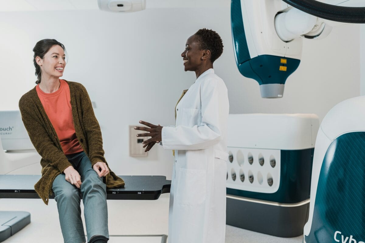 A woman in a white lab coat talking to another woman in a cardigan who is sat down, surrounded by medical equipment